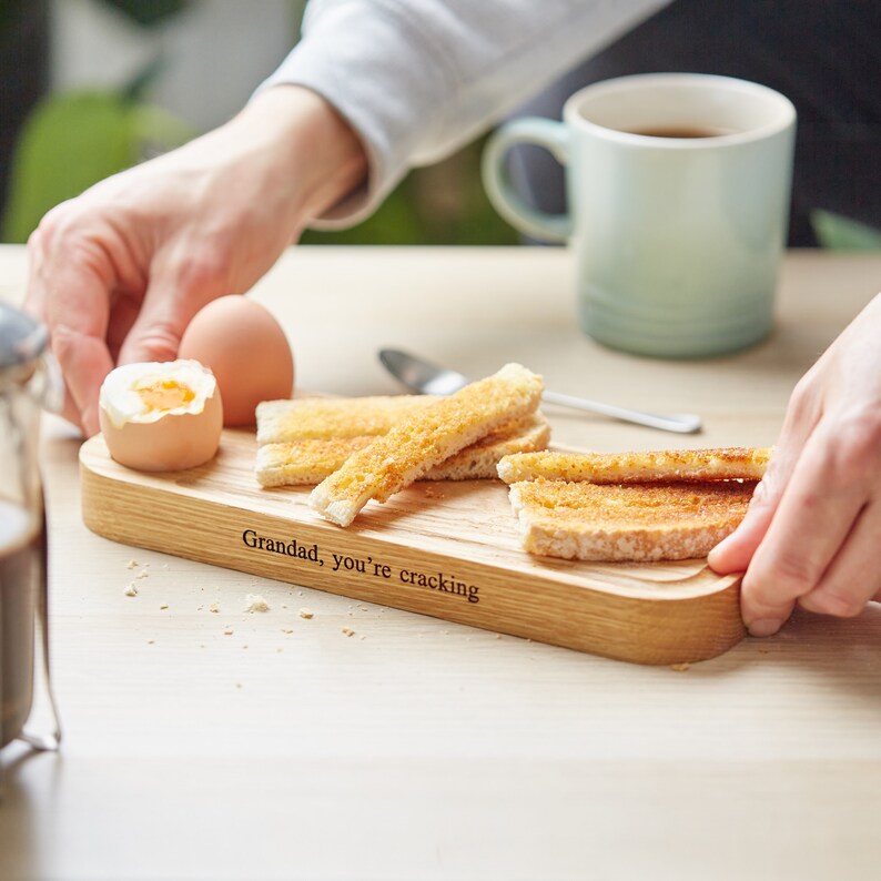 May include: A wooden serving board with the text "Grandad, you're cracking" engraved on it. The board is filled with toast and a boiled egg.