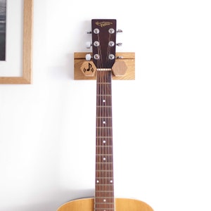 May include: A wooden guitar wall mount with a guitar hanging on it. The mount has two hexagonal wooden knobs with a music note and a treble clef carved into them. The guitar is a brown acoustic guitar with a wooden body and a brown neck.