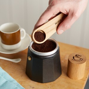 Może przedstawiać: Czarny ekspres do kawy jest napełniany zmieloną kawą za pomocą drewnianej miarki. Na miarce widnieje napis "PROPER COFFEE". Widoczny jest r&oacute;wnież kubek do kawy i drewniana pokrywka z motywem filiżanki.
