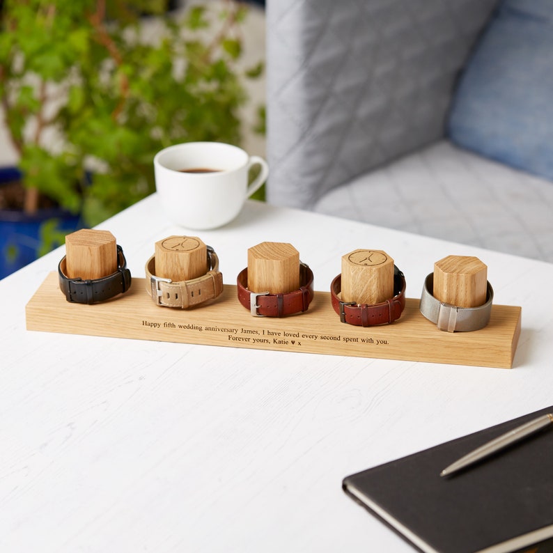 Op de afbeelding: Een houten horlogestandaard met vijf horloges. De horlogebandjes zijn zwart, beige, bruin en zilver. Elk horloge staat op een houten blok. De standaard is gegraveerd met de tekst: "Happy fifth wedding anniversary James, I have loved every second spent with you. Forever yours, Katie."