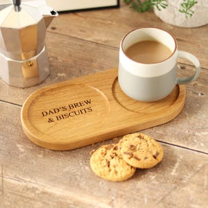 Può includere: Un vassoio di legno con una tazza e due biscotti. La tazza è grigia e bianca, piena di un liquido marrone chiaro. Il vassoio ha la scritta "DAD'S BREW & BISCUITS".