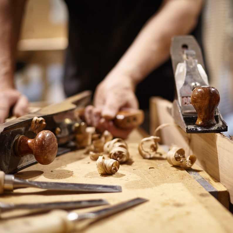 Op de afbeelding: Een houtbewerkingssc&egrave;ne met verschillende gereedschappen, waaronder schaven en beitels. Houtsnippers liggen verspreid op de houten werkbank. De gereedschappen hebben houten handvatten en metalen bladen. De afbeelding toont het houtbewerkingsproces.