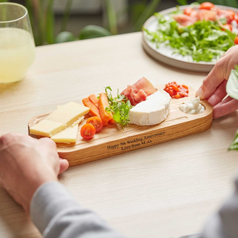 May include: A wooden serving board with a personalized message: "Happy 5th Wedding Anniversary Love from M...xx". The board is filled with cheese, crackers, fruit, and a small bowl of dip.