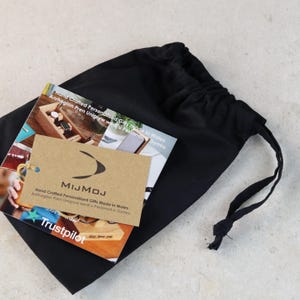 May include: A brown paper bag with the text "MIJMOJ Hand Crafted Personalised Gifts Made in Wales Anrhegion Pren Unigryw wedi'u Personoli o Gymru" printed on it. The bag is sitting on a black drawstring bag.