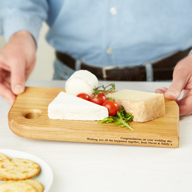 May include: A wooden cheese board with a selection of cheeses, cherry tomatoes, and arugula. The board has a rounded edge and a small circular indentation. Text on the board reads, "Wishing you all the happiness together, from David & Emily x. Congratulations on your wedding day."