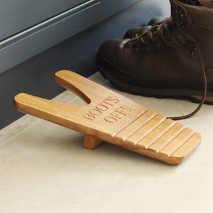 Pu&ograve; includere: Un cavastivali in legno marrone chiaro con la scritta "BOOTS OFF!" incisa sulla parte superiore. Il cavastivali ha una fessura per il tallone di uno stivale. Uno stivale in pelle marrone &egrave; sullo sfondo.