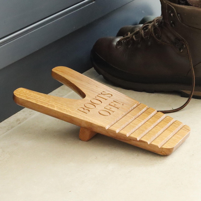 Pu&ograve; includere: Un cavastivali in legno marrone chiaro con la scritta "BOOTS OFF!" incisa sulla parte superiore. Il cavastivali ha una fessura per il tallone di uno stivale. Uno stivale in pelle marrone &egrave; sullo sfondo.