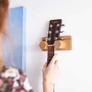 May include: A wall-mounted guitar hanger crafted from light-coloured oak wood, featuring two hexagonal supports that cradle the neck of a dark brown acoustic guitar. The headstock displays the text "Handmade by Hoku" in gold lettering.