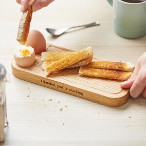 May include: A wooden serving tray with a  "Granddad, you're cracking" inscription. The tray has a small indentation for a boiled egg and is filled with slices of toasted bread.