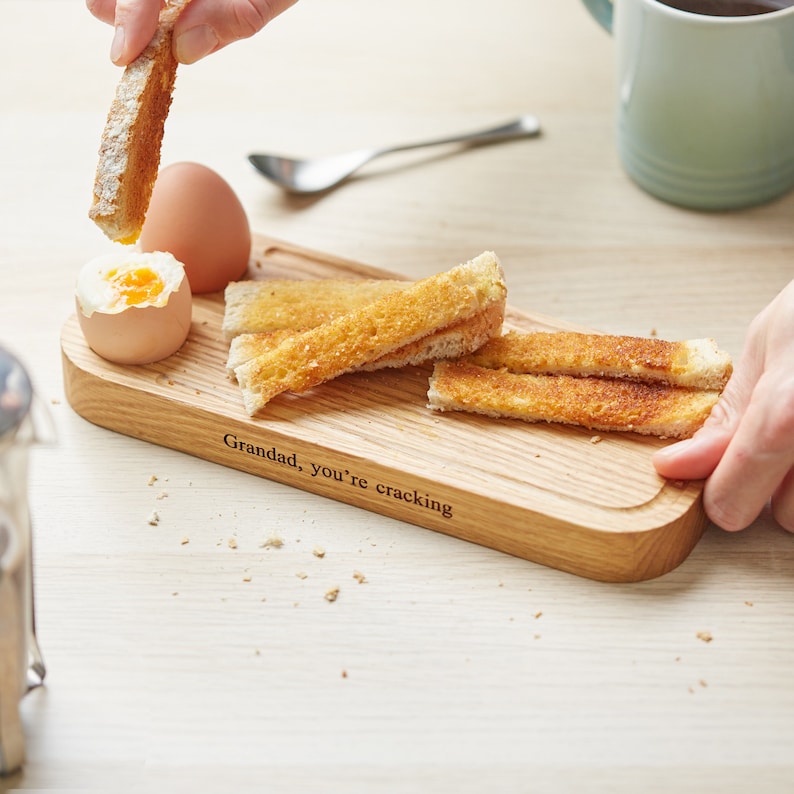 May include: A wooden serving tray with a  "Granddad, you're cracking" inscription. The tray has a small indentation for a boiled egg and is filled with slices of toasted bread.