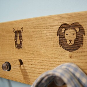 Op de afbeelding: Houten wandkapstok met een neushoorn- en leeuwenontwerp. De kapstok is gemaakt van lichtgekleurd hout en heeft drie haken. Een blauw-wit geruit item hangt aan een van de haken. Geschikt voor een kinderkamer.