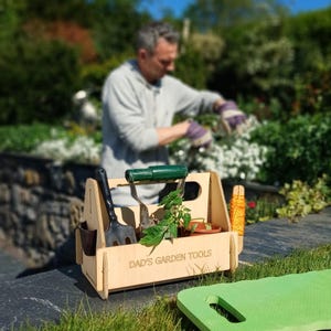 May include: A wooden garden tool caddy with the text "DAD'S GARDEN TOOLS" engraved on the front. The caddy holds a trowel, hand rake, and small potted plant. A person is gardening in the background.