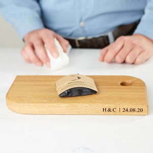 May include: A light-colored wooden cutting board with a rounded edge and a circular cutout. A small package with the text "MiJMoJ" is on the board. The text "H&C | 24.08.20" is also visible. A person is in the background.