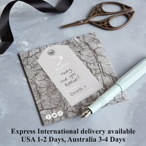May include: A light blue fountain pen rests on a gray tag with the text "Harry love you forever!" and a signature. A pair of decorative scissors and a black ribbon are also in the image. The tag has a tree bark texture.