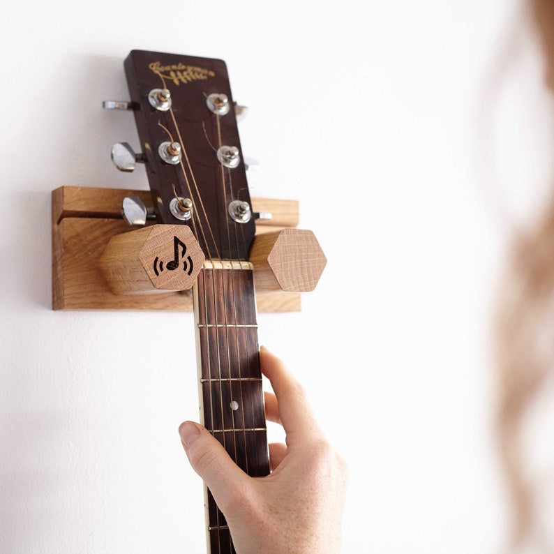 May include: Wooden guitar wall mount with a natural finish. The mount has hexagonal supports and a laser-etched musical note symbol. The guitar's headstock is visible, with tuning pegs and strings. A hand is reaching for the guitar.