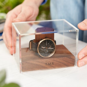 May include: A Skagen watch with a black face, gold-tone bezel, and black leather strap is presented in a clear acrylic display case. The watch sits on a wooden base with the initials "T.M.S" engraved. The case is made of clear acrylic.