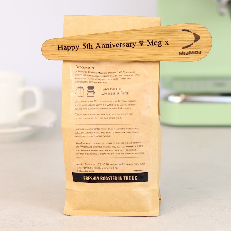 Może przedstawiać: Brązowa papierowa torba na kawę zamknięta drewnianym klipsem. Klips jest grawerowany napisem "Happy 5th Anniversary Meg x" i logo. Torba ma tekst, w tym "Freshly Roasted in the UK".