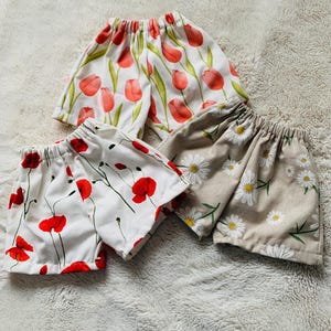May include: Three pairs of floral print shorts. One pair features red tulips on a white background, another has red poppies on white, and the third has white daisies on a tan background. The shorts have elastic waistbands.