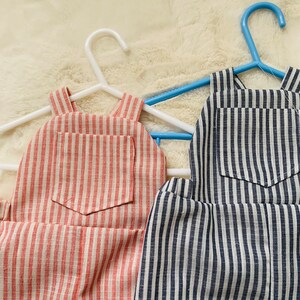 K&ouml;nnte beinhalten: Zwei Paar gestreifte Latzhosen h&auml;ngen an Kleiderb&uuml;geln. Ein Paar ist rot-wei&szlig; gestreift, das andere ist marineblau-wei&szlig; gestreift.