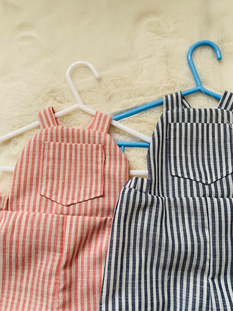 K&ouml;nnte beinhalten: Zwei Paar gestreifte Latzhosen h&auml;ngen an Kleiderb&uuml;geln. Ein Paar ist rot-wei&szlig; gestreift, das andere ist marineblau-wei&szlig; gestreift.