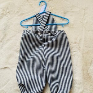 K&ouml;nnte beinhalten: Blau-wei&szlig; gestreifter Baby-Overall mit Kreuzriemen-Design und wei&szlig;en Kn&ouml;pfen.