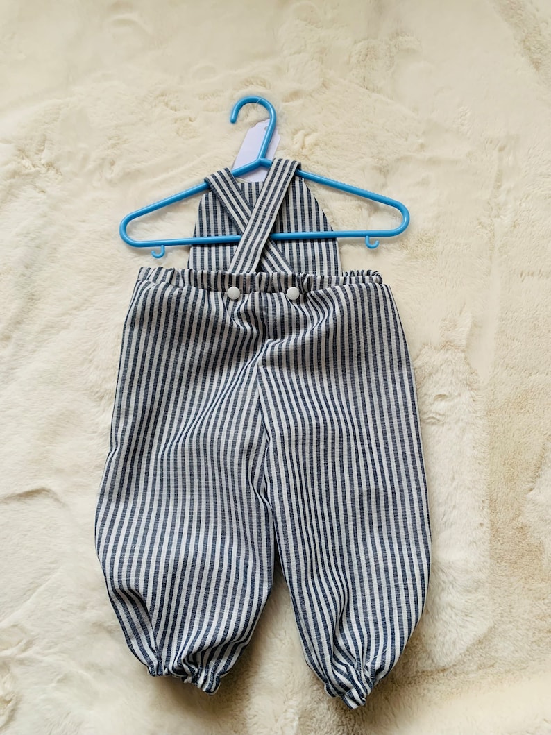 K&ouml;nnte beinhalten: Blau-wei&szlig; gestreifter Baby-Overall mit Kreuzriemen-Design und wei&szlig;en Kn&ouml;pfen.