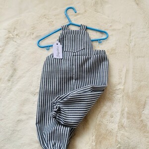 K&ouml;nnte beinhalten: Blaue und wei&szlig;e gestreifte Baby-Latzhosen mit einer kleinen Tasche auf der Vorderseite. Die Latzhosen h&auml;ngen an einem blauen Plastikb&uuml;gel mit einem Etikett, auf dem "Babyfashion" steht.