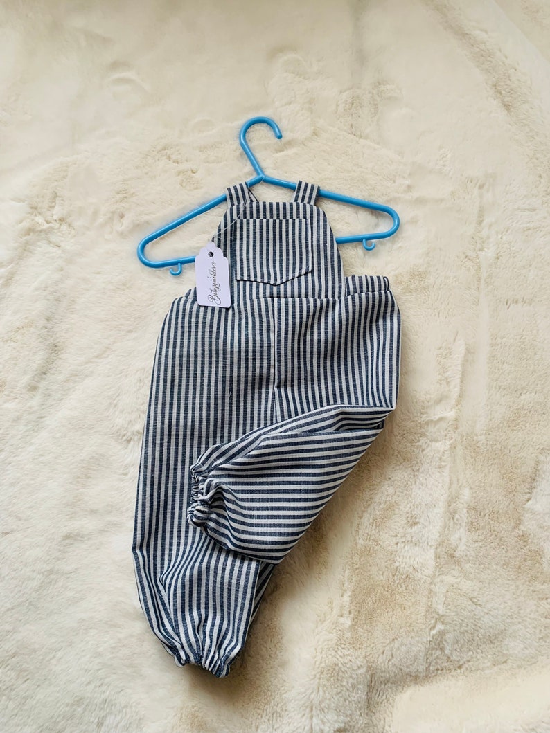 K&ouml;nnte beinhalten: Blaue und wei&szlig;e gestreifte Baby-Latzhosen mit einer kleinen Tasche auf der Vorderseite. Die Latzhosen h&auml;ngen an einem blauen Plastikb&uuml;gel mit einem Etikett, auf dem "Babyfashion" steht.