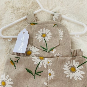 May include: Beige linen overalls with a daisy print. The overalls have a small pocket on the front and a tag that says "Baby Grace".
