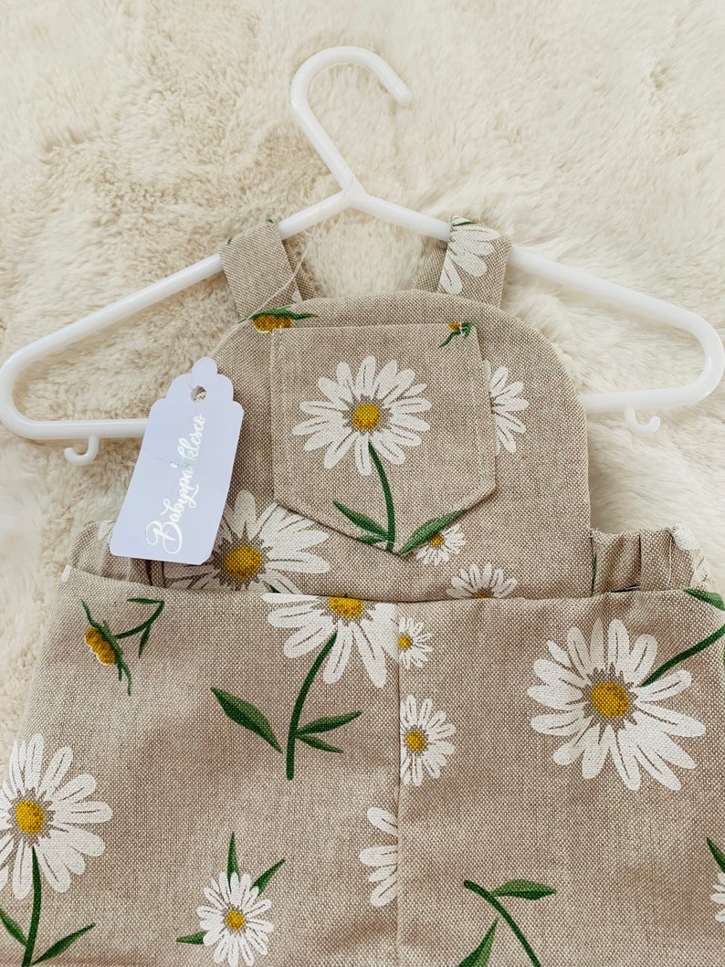 May include: Beige linen overalls with a daisy print. The overalls have a small pocket on the front and a tag that says "Baby Grace".