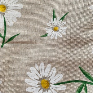 May include: A close-up of a beige fabric with a white daisy pattern. The daisies have yellow centers and green stems.