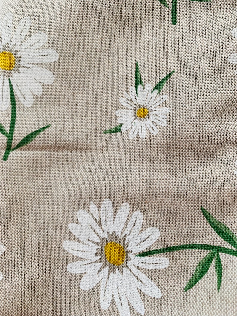 May include: A close-up of a beige fabric with a white daisy pattern. The daisies have yellow centers and green stems.
