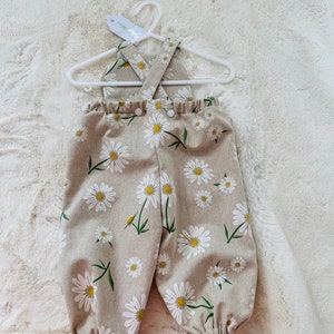 May include: Beige baby overalls with a white daisy print and criss-cross straps. The overalls have a button closure at the front.