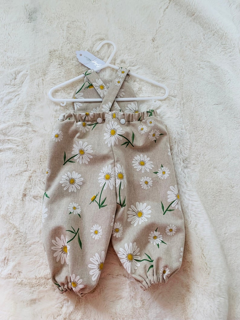 May include: Beige baby overalls with a white daisy print and criss-cross straps. The overalls have a button closure at the front.
