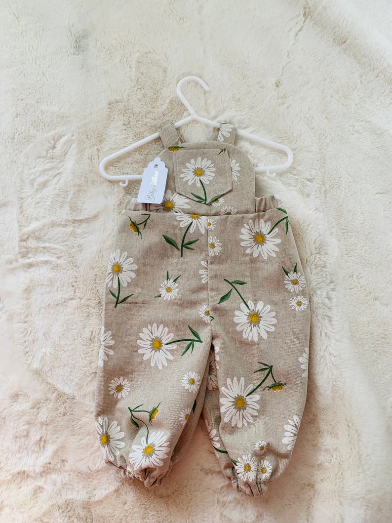 May include: A pair of beige linen overalls with a white daisy print. The overalls have a small pocket on the front and are hanging on a white hanger. The tag on the overalls says "Baby Shop".