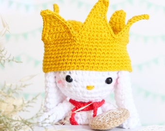 May include: A handmade crochet bunny doll with a yellow crown, red scarf, and a small, round, woven object. The bunny is white with black eyes and a yellow beak. The background is a light color with a decorative pattern.