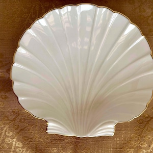 May include: A white, shell-shaped decorative dish with a gold trim. The dish has a glossy finish and a scalloped edge. The dish is likely made of ceramic or porcelain and is designed for decorative purposes.