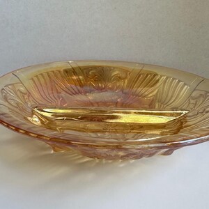 May include: An iridescent, oval glass dish with a central, rectangular insert. The dish features a decorative, embossed pattern and is mainly gold with pink and purple hues. It has small feet and handles.