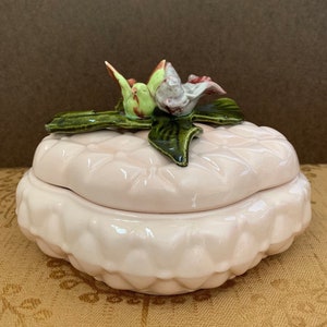 May include: A pink ceramic trinket box with a quilted lid and a ruffled base. The lid is adorned with a cluster of green leaves, a pink and white flower, and a small bird figurine. The box is a decorative item.