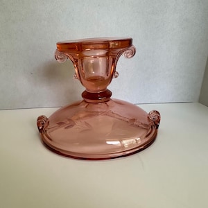 May include: Pink glass pedestal bowl with etched floral design. The bowl has a round base with two scrolled handles, a central stem, and a square top with scrolled accents. The glass has a translucent quality.