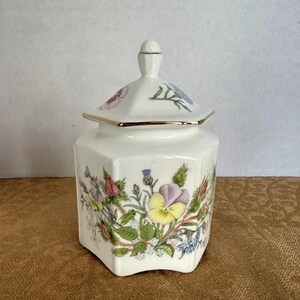 Peut inclure: Un pot en céramique blanc avec un couvercle, orné d'un motif floral avec des fleurs violettes, jaunes et vertes. Le pot a une bordure dorée et une forme hexagonale. Le couvercle est également hexagonal avec un petit bouton sur le dessus.