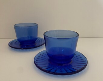 May include: Two cobalt blue glass teacups and saucers. The teacups are cylindrical with a slightly flared rim. The saucers are round with a ridged design. The set is displayed on a white surface.