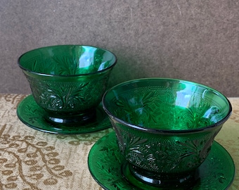 May include: Two emerald green glass bowls with matching saucers. The bowls and saucers have a detailed floral pattern. The bowls are round with a pedestal base. The saucers are flat and round.