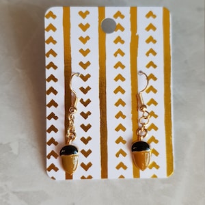Könnte beinhalten: Ein Paar goldene Eichel-Ohrringe mit schwarzen Oberteilen. Die Ohrringe werden auf einer weißen Karte mit einem goldenen Chevron-Muster präsentiert.