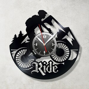Peut inclure: Horloge en vinyle noir avec une silhouette d'un cycliste de montagne sur un vélo avec le mot "Ride" en dessous. Le cadran de l'horloge est noir avec des chiffres et des aiguilles blancs.