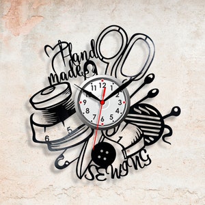 Puede incluir: Un reloj de vinilo en blanco y negro con temática de costura. La esfera del reloj es plateada con números y manecillas negros. El diseño incluye tijeras, hilo, botones y las palabras "Hand Made Sewing".