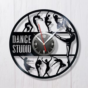 以下が含まれることがあります： 黒いビニールレコードの時計で、時計の文字盤と白い文字で「DANCE STUDIO」と書かれています。時計は、さまざまなポーズをとったダンサーのシルエットで飾られています。