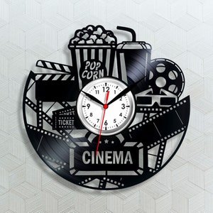 以下が含まれることがあります： 映画をテーマにした黒いビニールレコードの時計。時計には、フィルムリール、ポップコーンバケット、飲み物、3Dメガネ、クラッパーボードが描かれています。時計の文字盤は白で、黒い数字と針が付いています。時計は、「CINEMA」という文字が印刷された黒いビニールレコードで囲まれています。