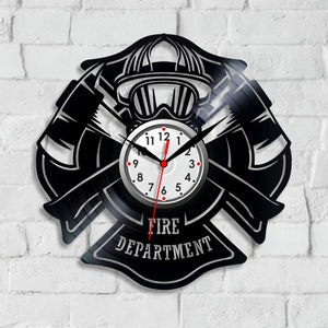 Puede incluir: Reloj de vinilo negro con un diseño de casco de bombero y hachas cruzadas. La esfera del reloj es blanca con números negros y manecillas rojas. El texto "FIRE DEPARTMENT" está impreso debajo de la esfera del reloj.