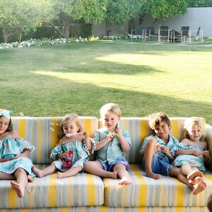 Puede incluir: Cinco ni&ntilde;os sentados en un sof&aacute; exterior a rayas amarillas y blancas. Llevan vestidos y camisas a juego azul y verde azulado con ribete de pomp&oacute;n blanco. Burbujas flotan en el aire. El fondo presenta un c&eacute;sped verde y &aacute;rboles.
